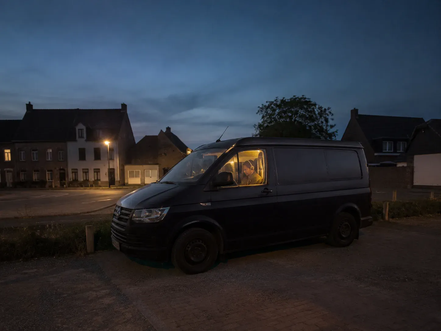 Zwart busje geparkeerd in de avond, verlichte cabine met persoon, huizen op de achtergrond.