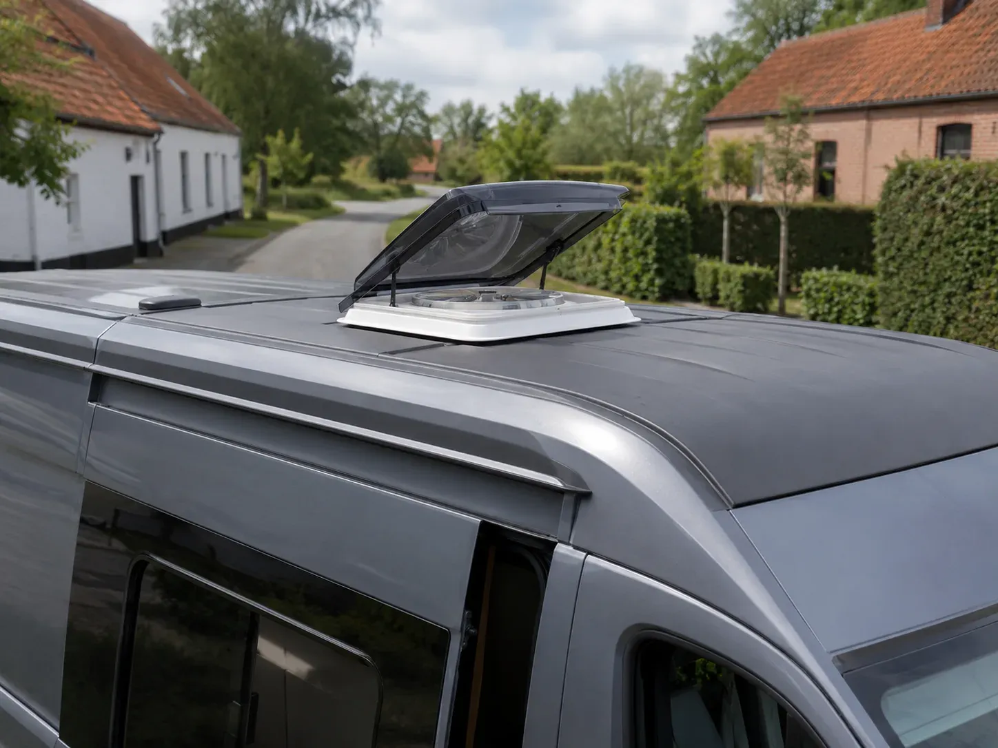 Grijze van met open dakluik voor ventilatie, geparkeerd in een landelijke omgeving.
