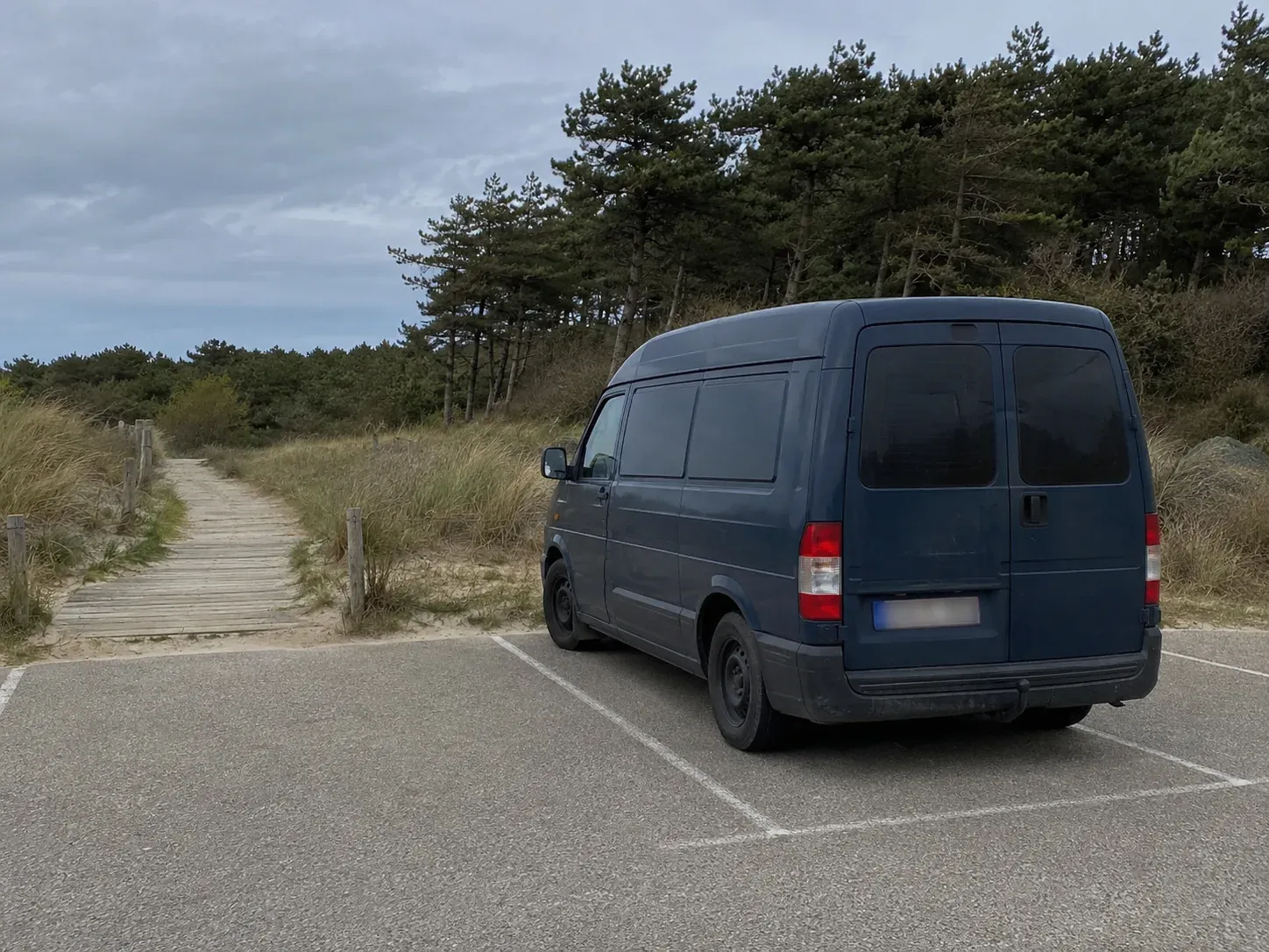 Donkerblauw busje geparkeerd bij houten pad in duinen met dennenbomen onder bewolkte lucht.