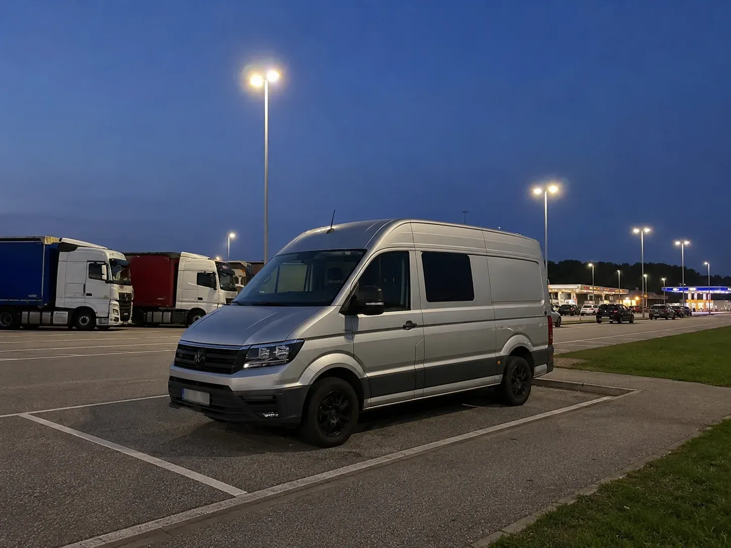 Zilveren van op een snelwegparking bij avond, met vrachtwagens en een verlicht tankstation in de verte.